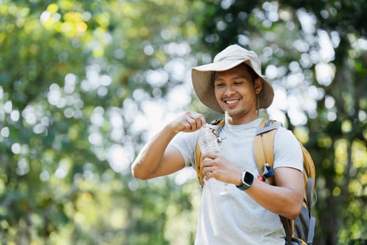 A young man with a backpack and hat enjoys a travel camping adventure in a lush green forest, smiling as he drinks from a water bottle on a sunny day.