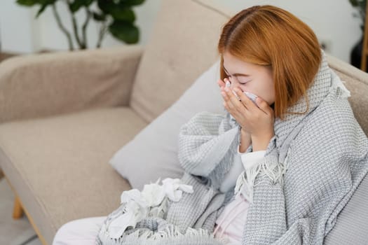 Woman feeling unwell, wrapped in a blanket, sneezing into a tissue while sitting on a couch at home.