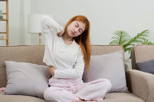Young woman feeling unwell at home, sitting on a couch with back pain, wearing comfortable loungewear in a modern living room.