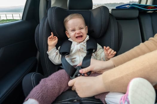 Crying toddler girl sitting in safety car seat. Baby is sad, upset and scared. Mom puts her baby into the carseat