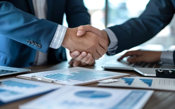 Two professionals shaking hands over a desk with documents and laptops, signifying a successful business deal.