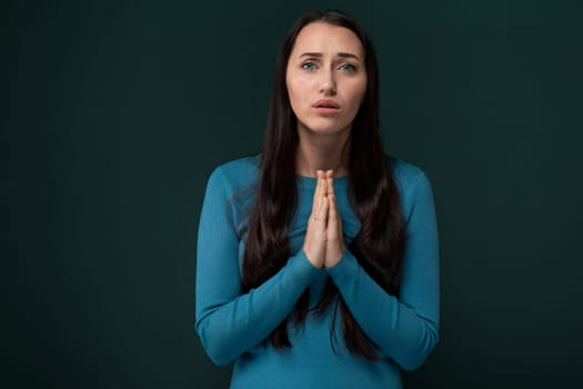 A woman wearing a blue shirt is shown praying in a kneeling position. Her hands are clasped together in front of her with eyes closed in concentration.