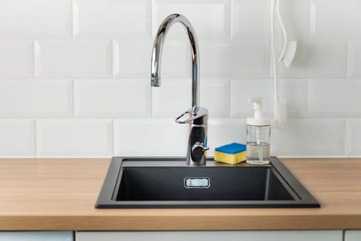 An empty black kitchen sink in a kitchen with a chrome faucet, wooden countertop, and white subway tile backsplash with dishwashing liquid, brush and sponge, banner with copy space