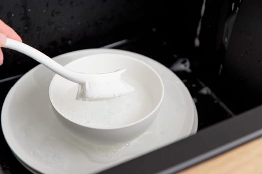 Washing of white plates dishes with brush in the black sink, tableware cleaning with soap foam concept