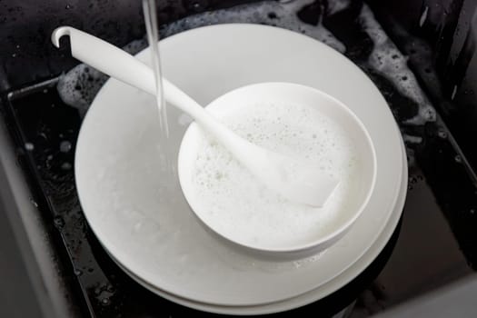 Washing of white plates dishes with brush in the black sink, tableware cleaning with soap foam concept