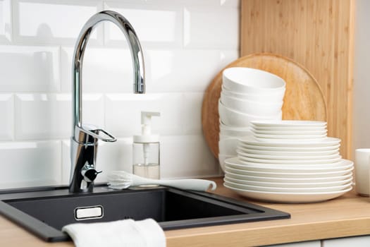 Clean washed tableware. Concept of washing dishes. Dishwashing liquid with brush, plates and cups near black kitchen sink with a chrome faucet on wooden counter and white subway tile backsplash