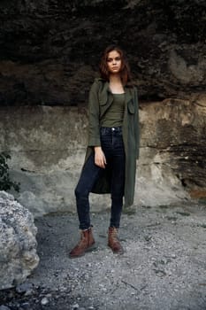 Stylish woman in green coat and jeans poses in front of rock wall with tree in background