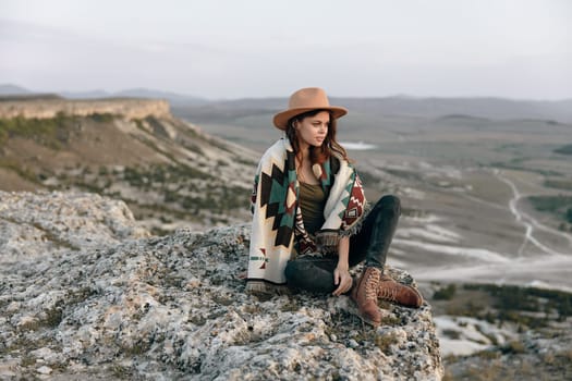 serene woman in the mountains sitting on a rock with a hat and blanket on her shoulders