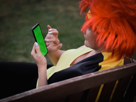 Portrait middle aged caucasian woman with Belgian flag wig with red hair on her head holding a mobile phone with green screen poking her finger while sitting on a bench in the backyard,side view closeup.Belgium day concept,flags,symbols,celebration,football,moms, fans, using technology, harvesting, using a smartphone, social media.