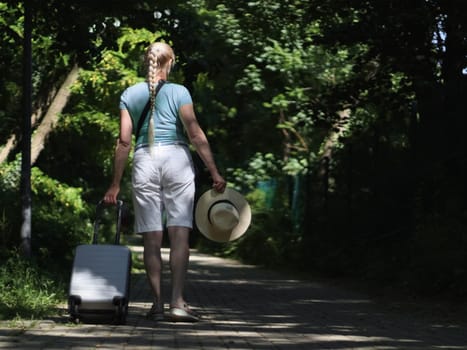 A young beautiful Caucasian blonde woman with a braided braid walks from behind with a suitcase and a straw hat in her hands on a summer sunny day along a path in the park, close-up side view.