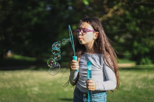 Portrait of one beautiful Caucasian brunette girl in glasses emotionally blowing a lot of soap bubbles, standing in the park on the playground, close-up view from below. Concept of PARKS and RECREATION, happy childhood, children's picnic, happy childhood, outdoor recreation, playgrounds.