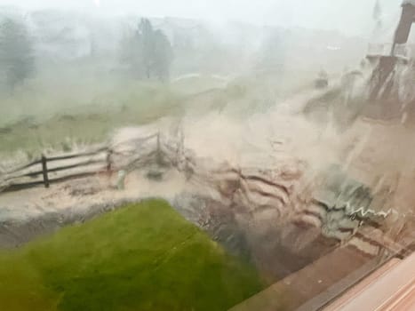 A view through a rain-streaked window capturing a heavy downpour with hail and flash flooding in a suburban backyard. The scene shows blurred fences, trees, and lawn due to the intense rain and hail.