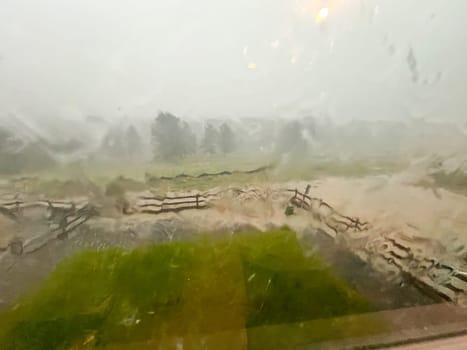 A view through a rain-streaked window capturing a heavy downpour with hail and flash flooding in a suburban backyard. The scene shows blurred fences, trees, and lawn due to the intense rain and hail.