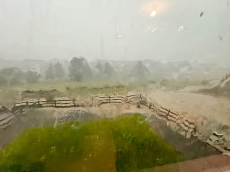 A view through a rain-streaked window capturing a heavy downpour with hail and flash flooding in a suburban backyard. The scene shows blurred fences, trees, and lawn due to the intense rain and hail.