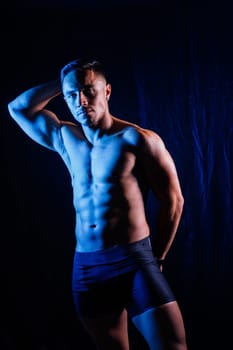 A man with a bare chest is posing against a dark backdrop, showing off his muscular physique. His head, hands, stomach, arms, shoulders, legs, and neck are visible in the flash photography