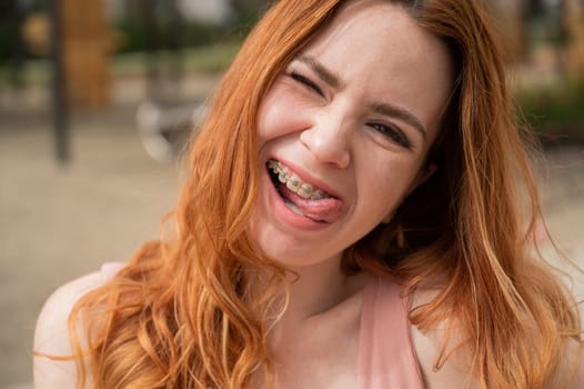 Young woman with braces on her teeth smiles and shows her tongue outdoors