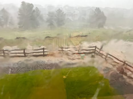 A view through a rain-streaked window capturing a heavy downpour with hail and flash flooding in a suburban backyard. The scene shows blurred fences, trees, and lawn due to the intense rain and hail.