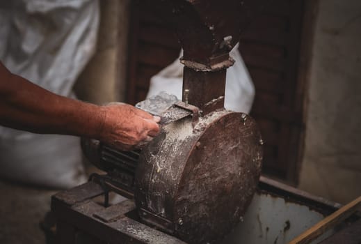 One elderly Caucasian unrecognizable man makes dry animal feed in a homemade crusher, pushing back the iron flap with his hand, standing on the street of a house in a village in the countryside, close-up side view from above.