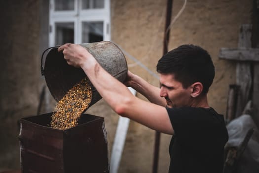 One young Caucasian brunette pours a mixture of wheat and corn from a bucket into a crusher for preparing dry food, standing on the street near a village house, close-up side view in dark style.