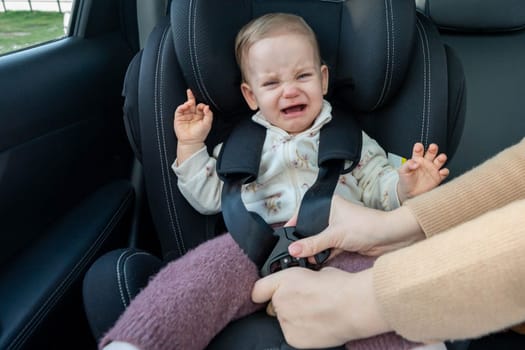 Mother puts her toddler baby into the car seat. Baby is sad and crying