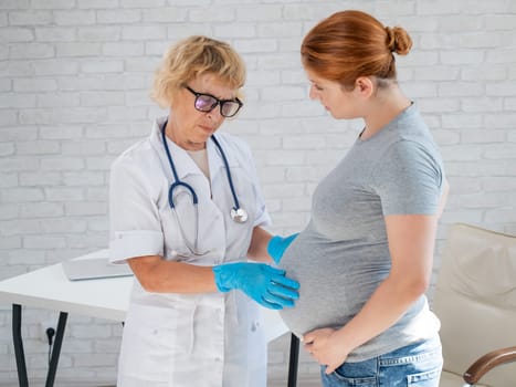 Pregnant woman visiting a doctor. Elderly Caucasian female gynecologist holds hands on the tummy of a pregnant patient