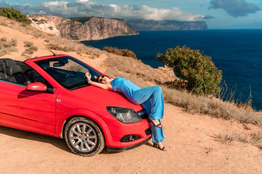 A woman is laying on top of a red convertible car. The car is parked on a dirt road near the ocean. The woman is wearing a blue dress and she is enjoying the moment