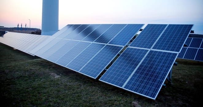 Rows of blue photovoltaic panels stand in an open field, capturing the last light as the sun sets on the horizon. The sky displays a gradient of colors from blue to soft orange, signifying the transition from day to night. The solar farm generates sustainable energy in a rural setting, highlighting the blend of technology with the natural environment.