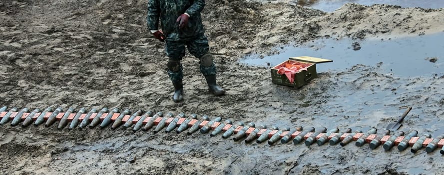 A soldier in uniform prepares weapons in a muddy field, surrounded by ammunition boxes. The low angle shot makes the soldier look powerful and intimidating, capturing the horror and futility of war.