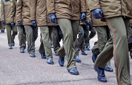 A group of soldiers dressed in green military uniforms march together in formation on a parade ground during a military ceremony. They are wearing black gloves and their faces are not visible.