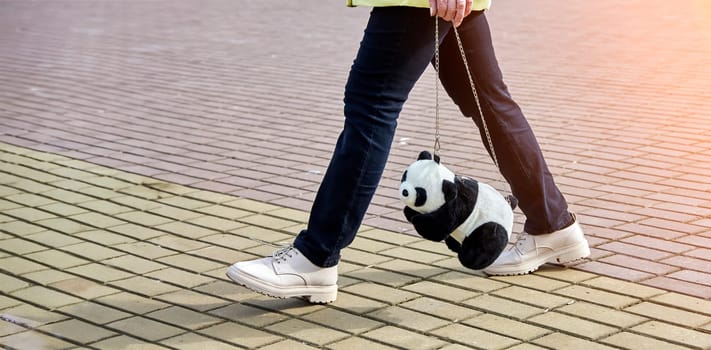 A fashionable young woman strolls with a cute panda plush bag, donning a chic ensemble and white kicks. The panda accessory brings a fun, whimsical touch to her urban style.