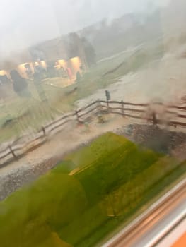 A view through a rain-streaked window capturing a heavy downpour with hail and flash flooding in a suburban backyard. The scene shows blurred fences, trees, and lawn due to the intense rain and hail.