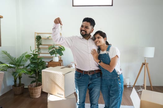 Indian couple moving into a new home, holding keys, surrounded by cardboard boxes and houseplants, celebrating new beginnings in a modern living room.