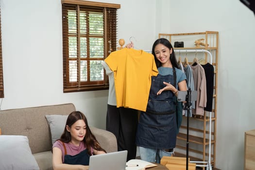 Two young women running an online e-business, showcasing clothing items, and engaging with customers through live streaming and laptop in a modern home office.