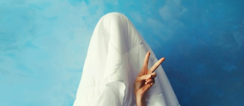 Cheerful lazy woman waking up after sleeping covered head with white blanket showing cool gesture her fingers on blue background