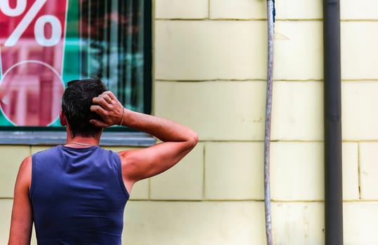 A man is standing outside a store, scratching his head as he observes a large percentage sign displayed in the window. His posture suggests he might be puzzled or considering the implications of the advertised discount, sale, or promotion.