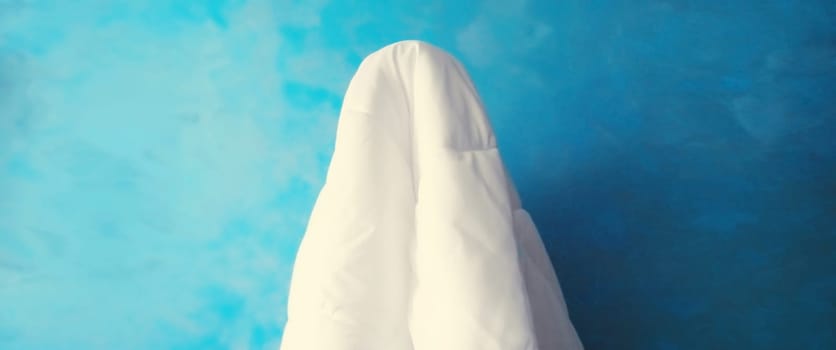 Cheerful lazy woman waking up after sleeping covered head with white blanket on blue background at home