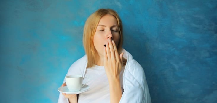 Sleepy lazy young woman yawns waking up after sleeping wrapped in white soft comfortable blanket holds cup of coffee in morning on blue background at home