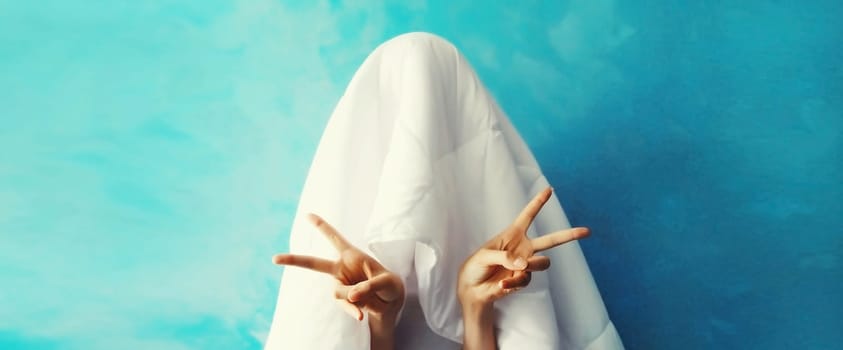 Cheerful lazy woman waking up after sleeping covered head with white blanket showing cool gesture her fingers on blue background