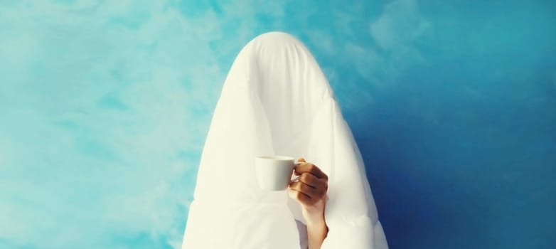 Cheerful lazy woman waking up after sleeping covered head with white soft blanket holds cup of coffee on blue background at home