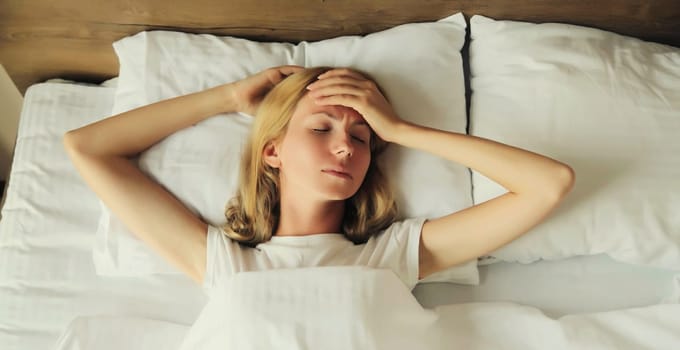 Upset exhausted woman with headache, pain tension in the head, suffers from migraine lying on the bed on pillow in bedroom at home