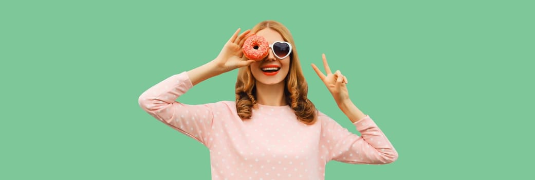 Portrait of happy cheerful laughing young woman with sweet donut having fun wearing heart shaped sunglasses on green background