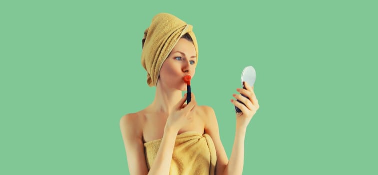 Portrait of beautiful young woman painting her lips with red lipstick applying makeup looking in pocket mirror and drying her wet hair with wrapped bath towel on her head on green background