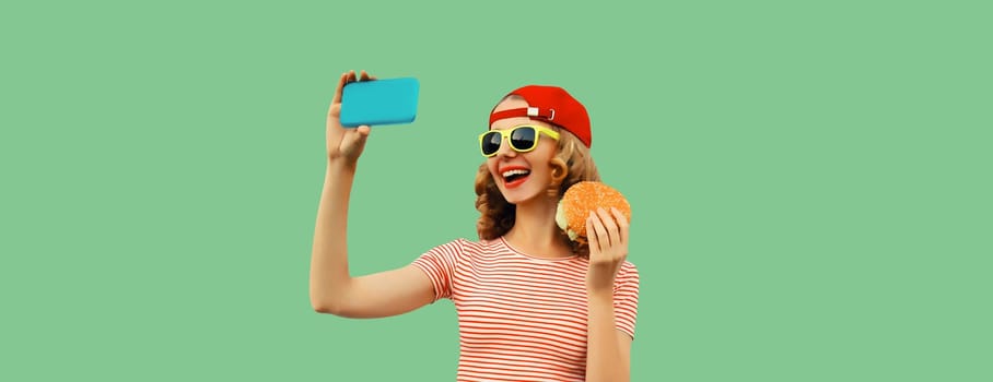 Portrait of modern cheerful laughing young woman taking selfie with smartphone and burger fast food on green studio background