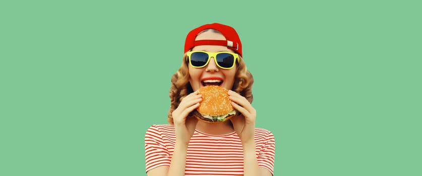 Portrait of stylish modern cheerful young woman eating burger fast food isolated on green studio background
