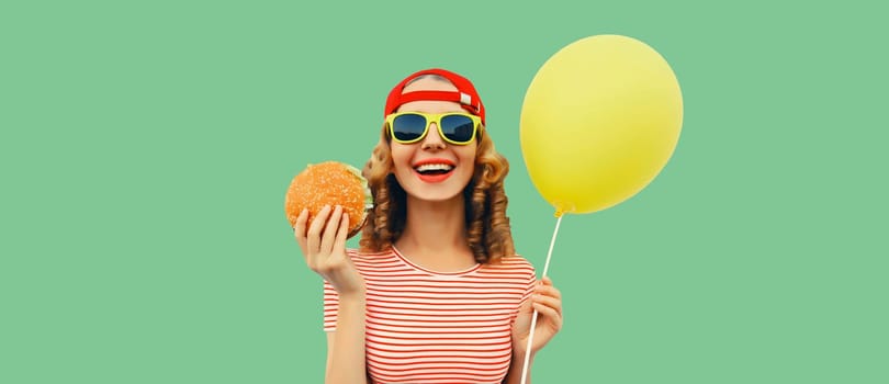 Portrait of cheerful funny young woman with burger fast food and yellow balloon on green studio background