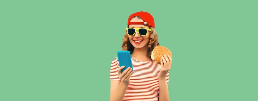 Portrait of stylish happy smiling young woman with mobile phone and burger fast food on green studio background