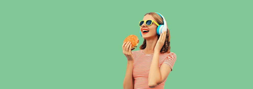 Portrait of happy laughing young woman listening to music in headphones holding burger fast food on green studio background, banner copy space for advertising text