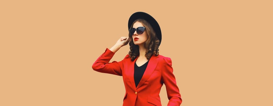 Stylish elegant woman posing in business suit, red blazer jacket and black round hat on brown studio background