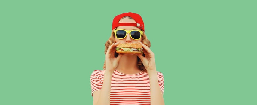 Portrait of stylish modern cheerful young woman eating burger fast food isolated on green studio background