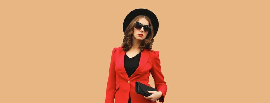 Stylish elegant woman posing in business suit, red blazer jacket, black round hat with handbag clutch posing on brown studio background